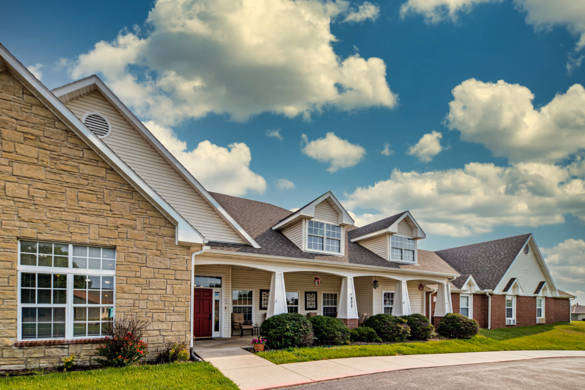 Exterior view of a senior living facility building with a stone and brick facade, beige siding, and a covered porch with chairs and hanging flower baskets under a partly cloudy blue sky.