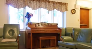 Small sitting room with an upright piano under a window flanked by green upholstered chairs.