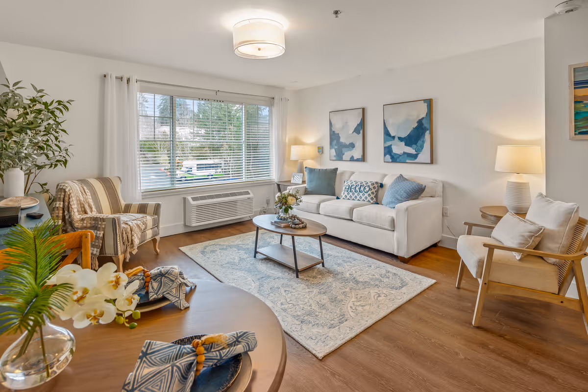 A bright and cozy living room with a large window covered by white curtains and blinds. The room features a white sofa with blue and patterned pillows, two armchairs, a wooden coffee table on a light blue patterned rug, and two table lamps on side tables. There are two abstract blue and white paintings on the wall above the sofa. In the foreground, a wooden dining table is partially visible with blue patterned napkins and a vase with white flowers and green leaves.