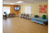 A spacious rehabilitation room with exercise equipment including a stationary bike, chairs lined up against the walls, a treatment table with a green exercise ball on it, a wall-mounted TV, and a colorful poster on the wall. The room has large windows with blinds and a light-colored floor.