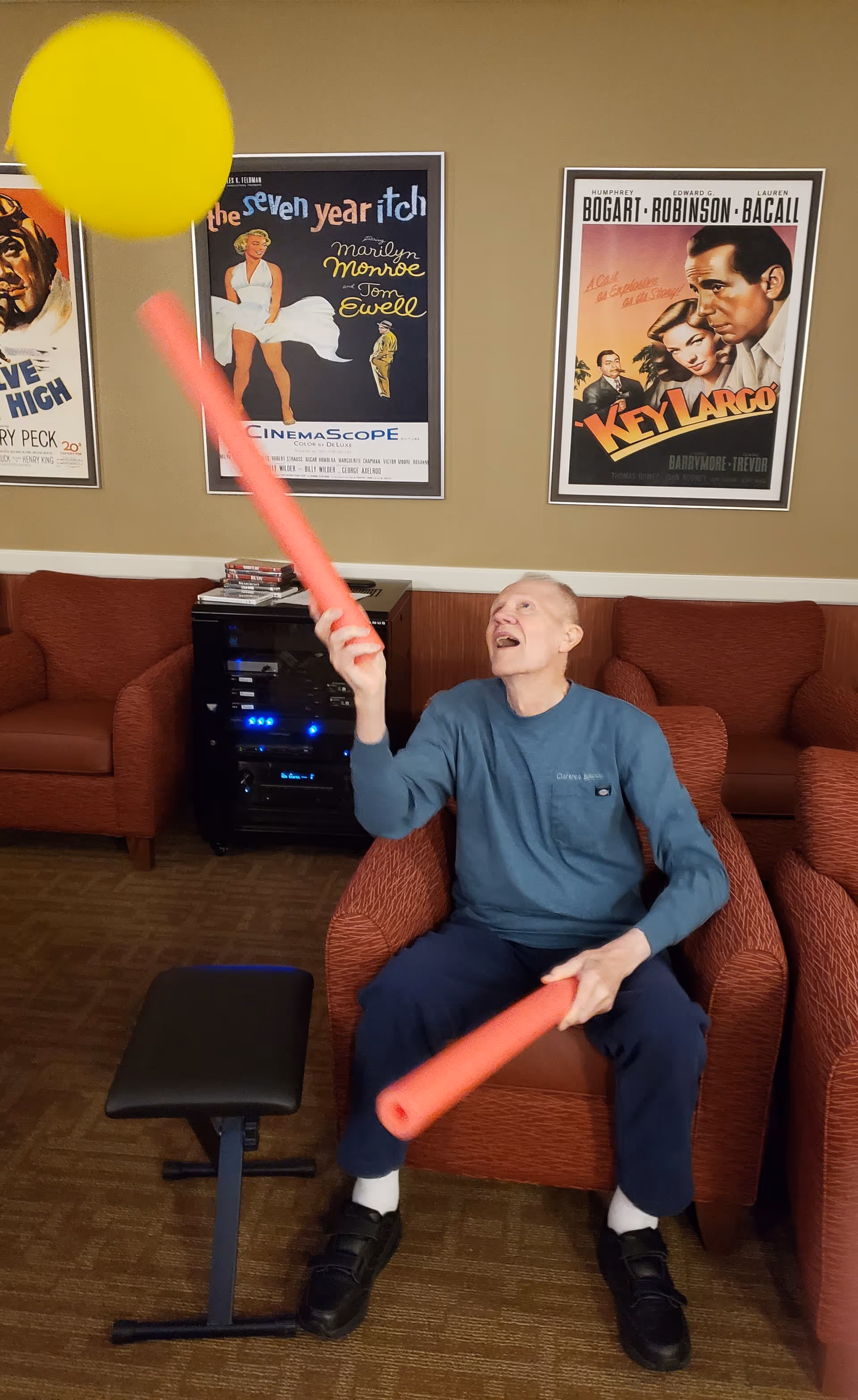 An elderly man sitting in a red armchair in a lounge area, playing with a yellow balloon using two red foam sticks. Behind him are three vintage movie posters on the wall and two more red armchairs. There is also a small black bench and an audio-visual equipment cabinet in the background.