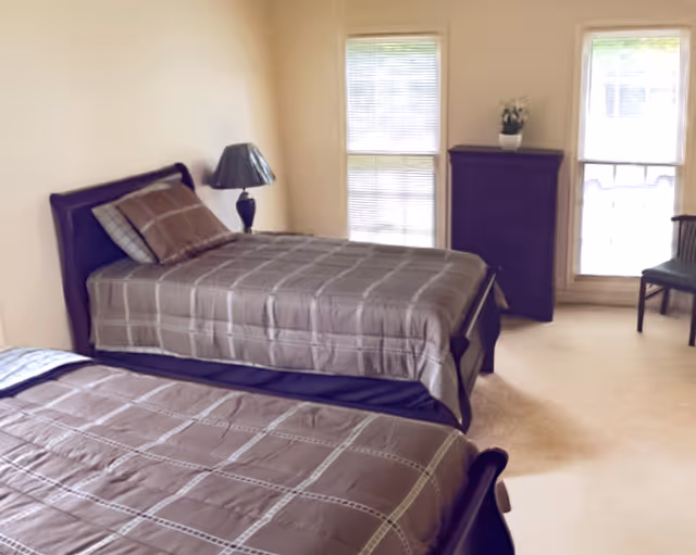 A bedroom with two single beds covered in matching brown plaid bedspreads. There is a bedside table with a lamp between the beds, a dark wooden dresser against the wall, and two large windows with blinds letting in natural light. A chair is placed near one of the windows.