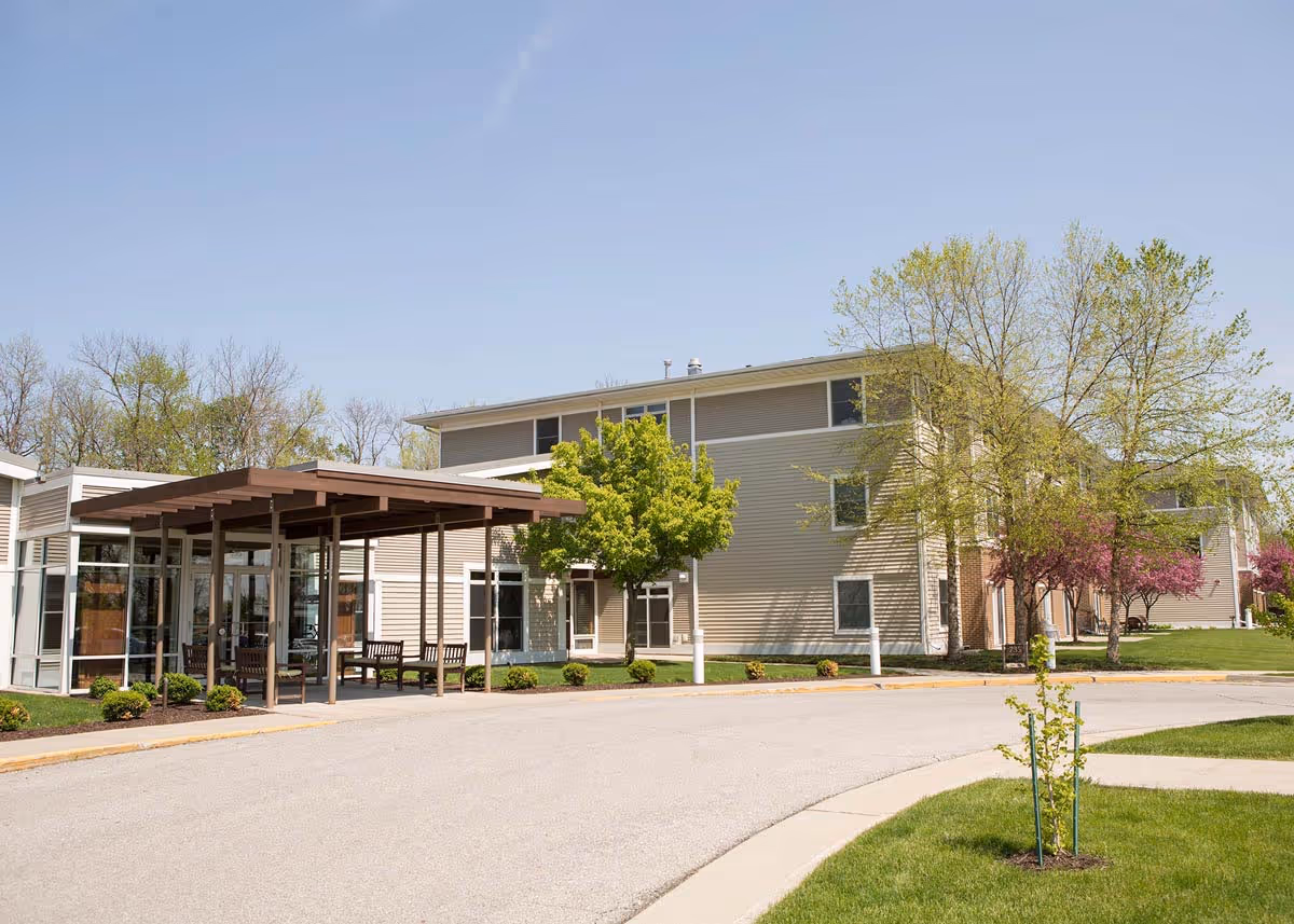 Exterior view of a senior living facility building with beige siding, large windows, a covered entrance with benches, and surrounding trees and greenery under a clear blue sky.