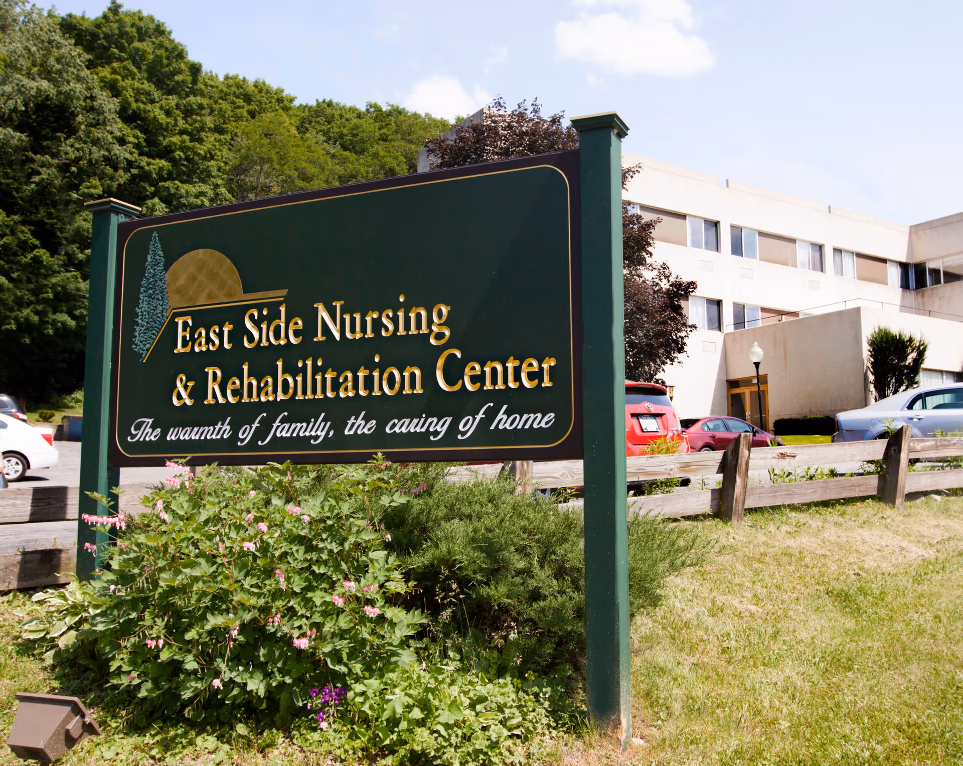 Outdoor view of the East Side Nursing & Rehabilitation Center sign with a building and parked cars in the background, surrounded by greenery and a wooden fence.