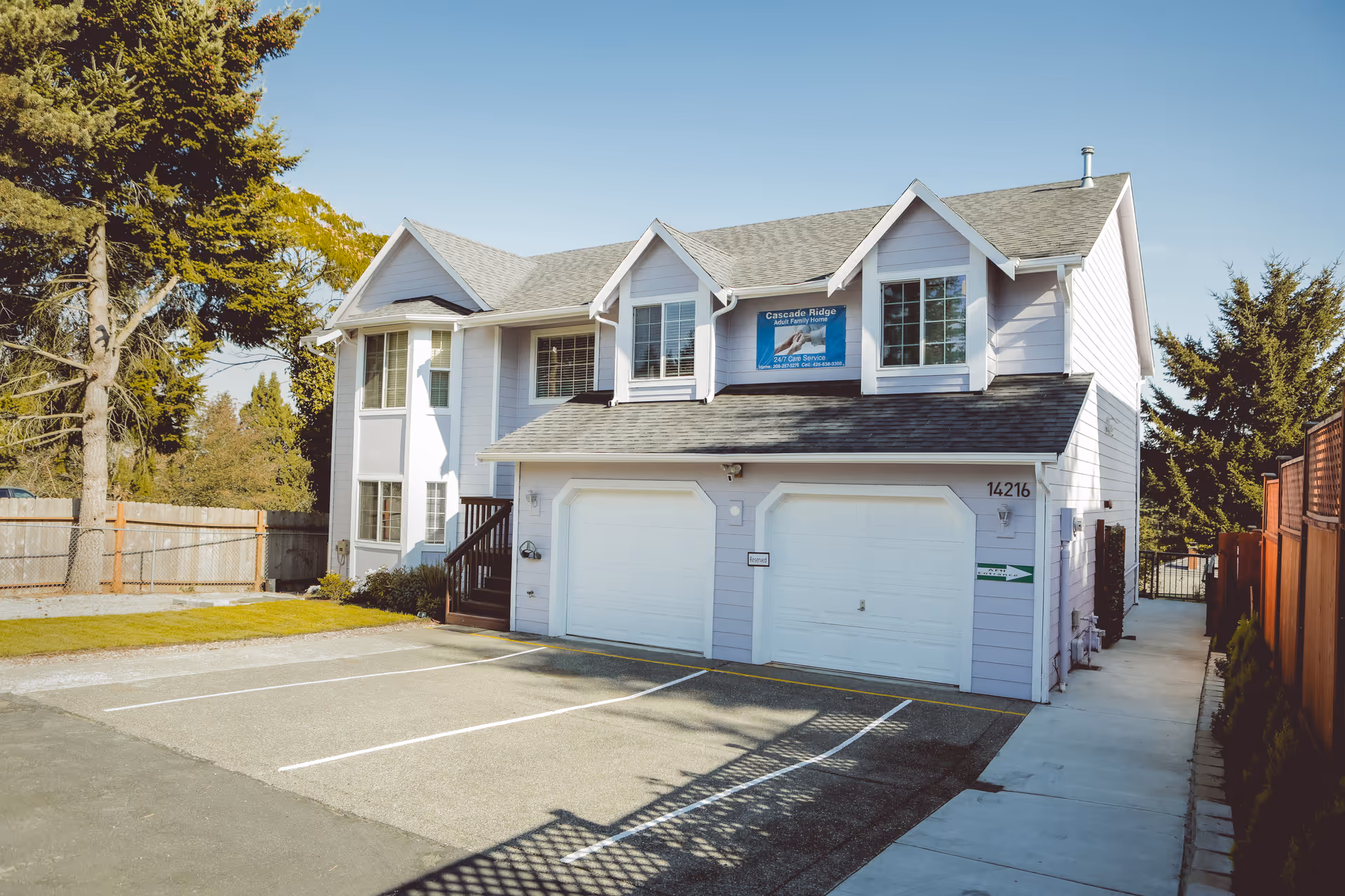 Exterior view of a two-story residential building with light purple siding and a gray shingled roof. The building has two white garage doors, a small staircase leading to the entrance, and a sign above the garage that reads 'Cascade Ridge Adult Family Home'. There is a paved parking area with marked spaces in front and trees and fencing surrounding the property.