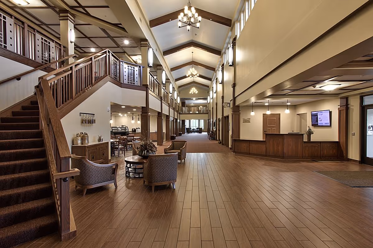 Spacious senior living facility lobby with high vaulted ceilings, wooden staircase on the left, seating area with chairs and a table, a reception desk on the right, and a dining area with tables and chairs in the background.