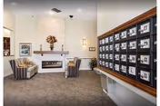 A cozy assisted living facility common area with a fireplace in the center, flanked by two striped armchairs and a small table with a lamp. On the right side, there is a large wooden mail organizer with numbered mail slots. The room has neutral-colored walls and carpeted flooring, with a decorative flower arrangement on the fireplace mantel.
