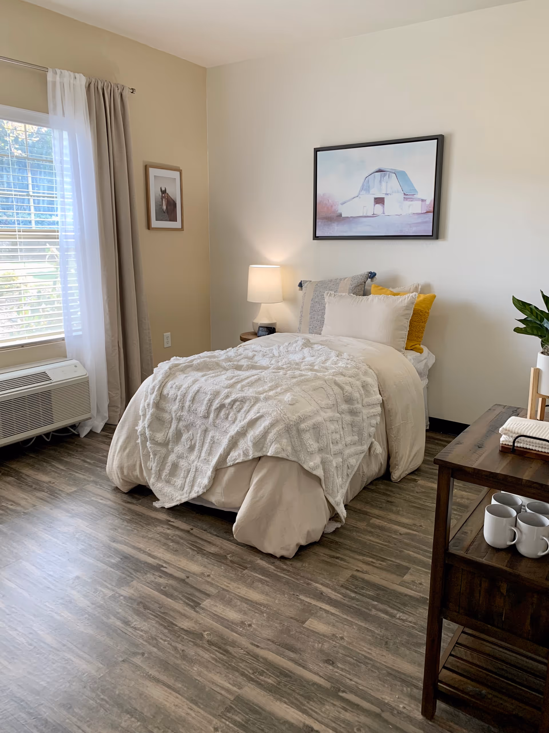 A cozy bedroom with a single bed covered in beige and white bedding, including a textured white blanket and multiple pillows. A bedside table with a lamp is next to the bed. The room has wood flooring, a window with beige and white curtains, and framed artwork on the walls. A wooden side table holds several white mugs and a potted plant.