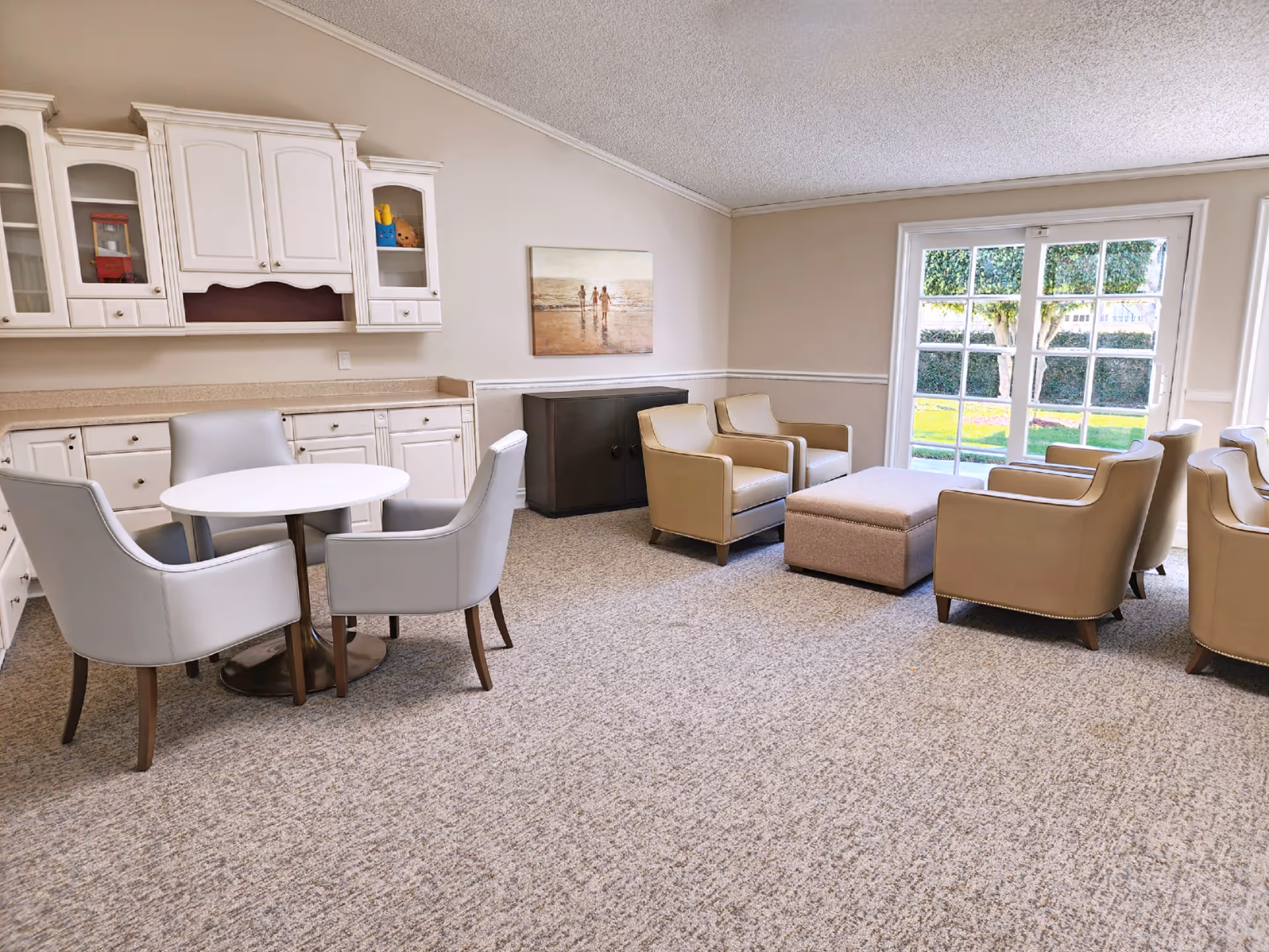 A bright and cozy common area with beige walls and carpeted floor. The room features a round white table surrounded by four gray upholstered chairs on the left side, white cabinetry with glass doors above the counter, and a seating area with four beige armchairs and a matching ottoman near a large window with a view of green trees and grass outside. A painting of people on a beach hangs on the wall above a dark wooden cabinet.
