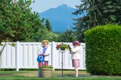 Two elderly women gardening outdoors near a white fence with a mountain and trees in the background. One woman is watering plants while the other is tending to a hanging flower basket.