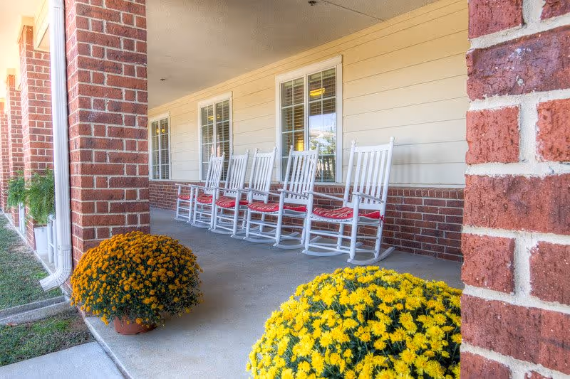 A covered porch area with a row of white rocking chairs with red cushions lined up against a beige wall with windows. There are two large pots of yellow and orange flowers on the concrete floor near the brick columns supporting the porch roof.