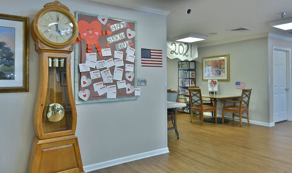 Interior view of a senior living facility showing a wooden grandfather clock on the left, a bulletin board with heart decorations and notes titled 'Staff Shout Out', an American flag on the wall, and a small dining area with a table and chairs. There is a bookshelf filled with books and games, a framed floral painting on the wall, and a hanging decoration with the year '2021'. The floor is wooden and the walls are painted light beige.