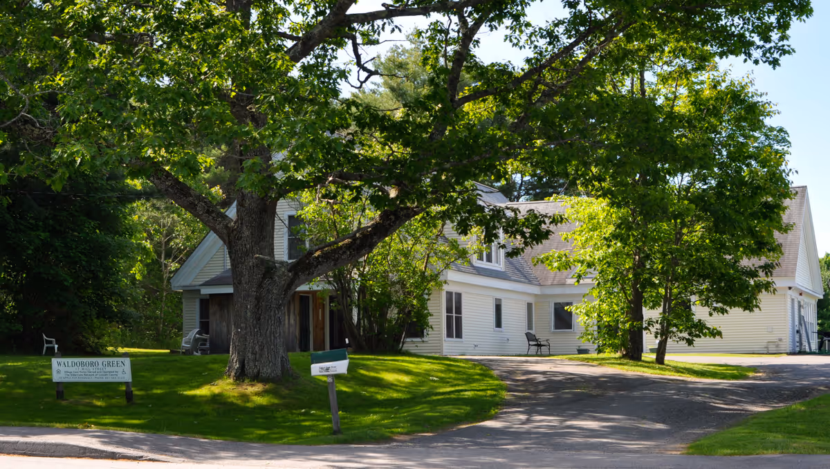 A white building with a sloped roof surrounded by green trees and grass. There is a large tree in the foreground and a paved driveway leading to the building. A sign near the tree reads 'Waldoboro Green'.