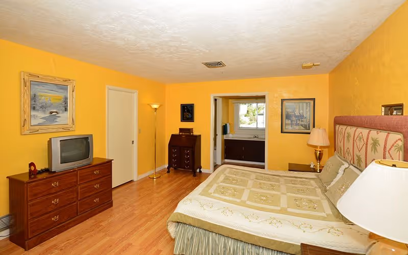 A bedroom with yellow walls and wooden flooring featuring a bed with a patterned headboard and beige bedding. There is a wooden dresser with a small TV on top, a floor lamp, a small chest of drawers, and a bedside table with a lamp. An open doorway leads to a bathroom with a window and a sink visible.