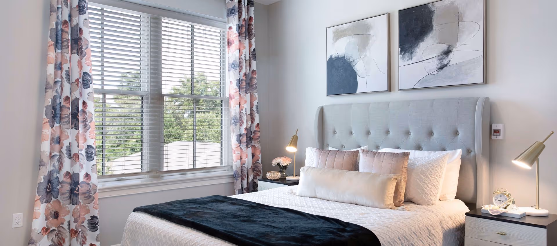 Bright bedroom with a tufted headboard, neatly made bed, floral curtains, bedside tables with lamps, and two abstract paintings above the bed.