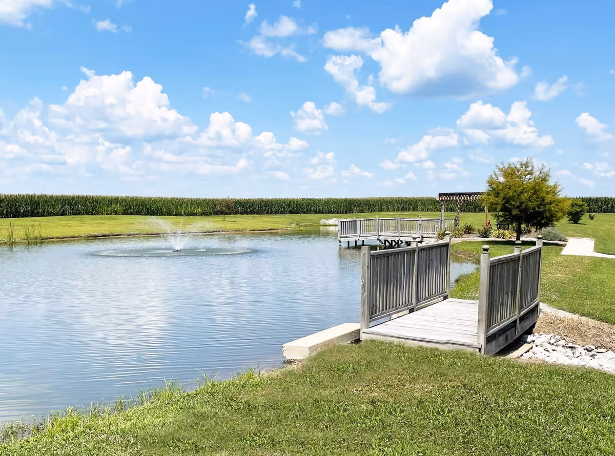 A peaceful outdoor scene at Courtyard Estates of Girard featuring a small pond with a water fountain, a wooden dock extending over the water, a grassy area, a tree, and a pergola in the background under a blue sky with scattered clouds.
