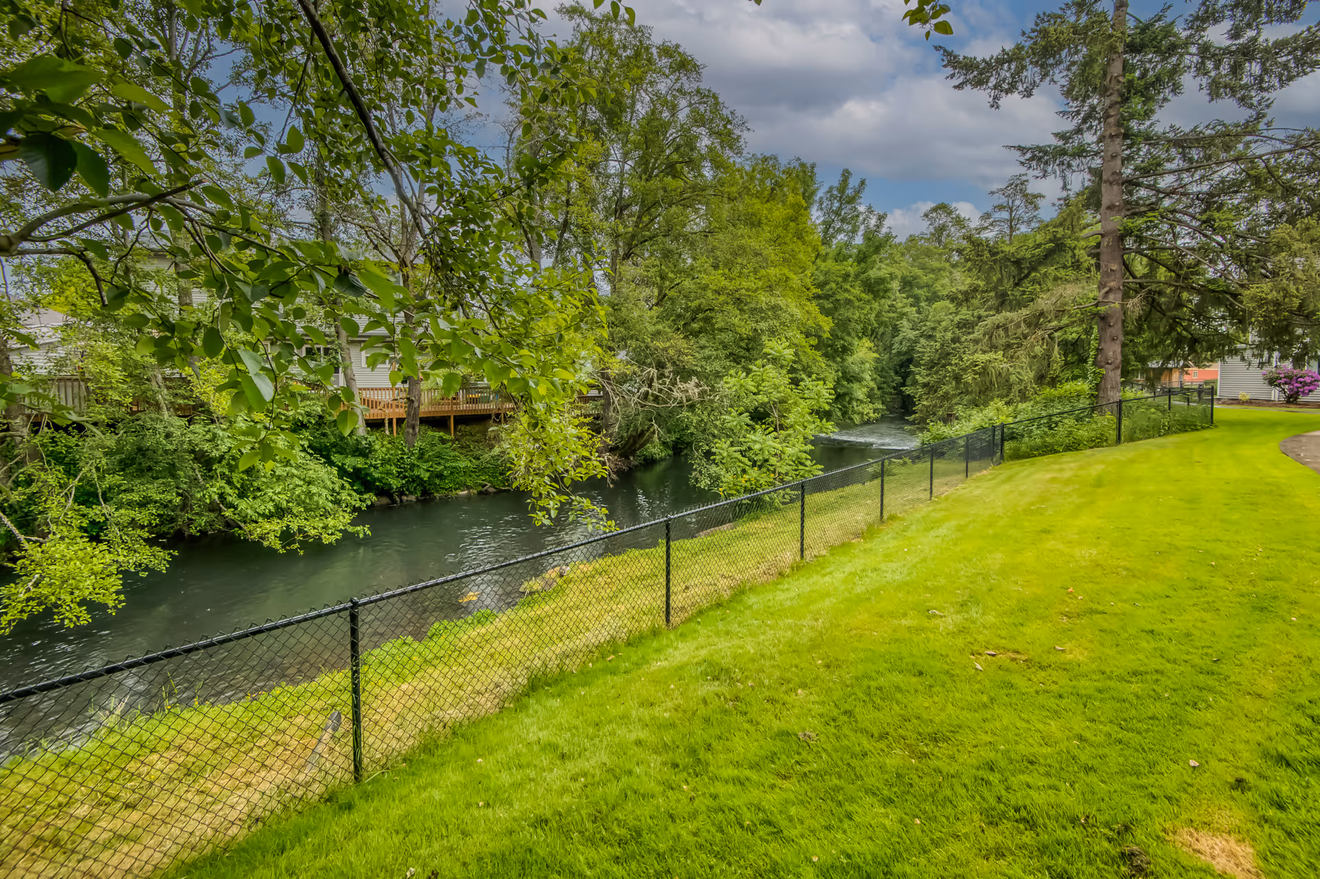 A lush green lawn bordered by a black chain-link fence next to a flowing river surrounded by dense trees and foliage under a partly cloudy sky.