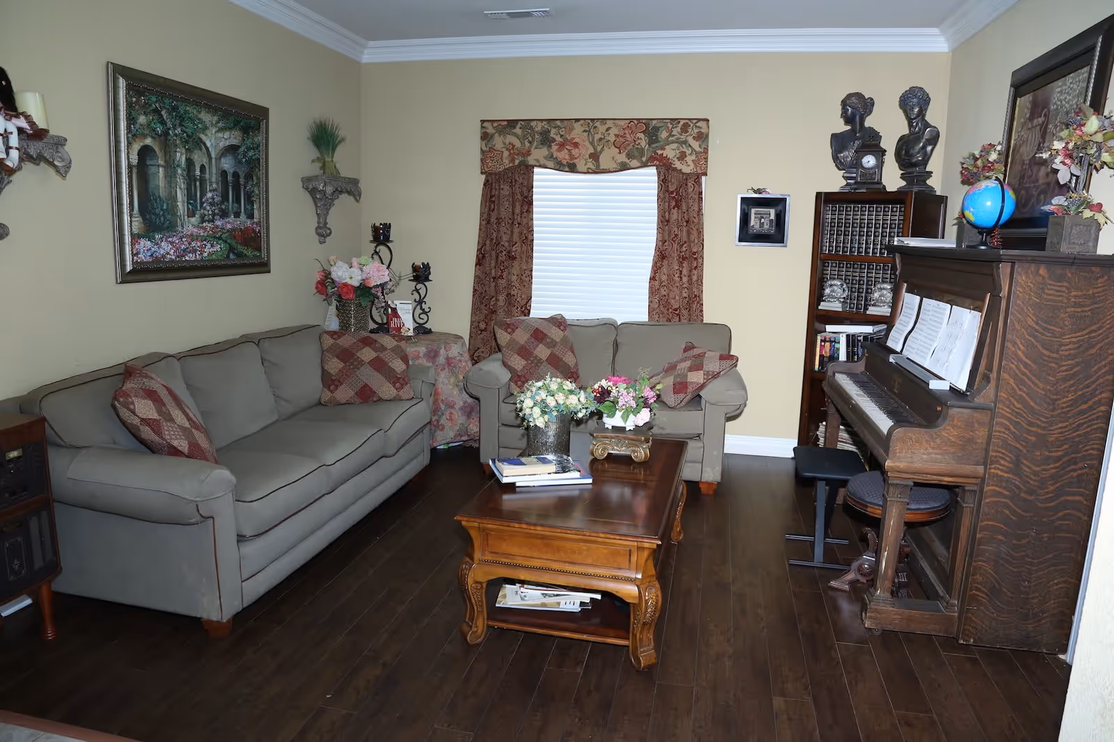 A cozy living room with two beige sofas adorned with patterned cushions, a wooden coffee table with flower arrangements and books, a wooden upright piano with sheet music, a bookshelf with decorative items and books, a window with floral curtains, and framed artwork on the walls.