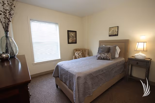 A cozy bedroom with a single bed covered in a gray quilt and multiple pillows. There is a wooden nightstand with a lamp and decorative item on the right side of the bed. A cushioned chair with a patterned pillow is placed in the corner near a window with closed blinds. A wooden dresser with a large glass vase and decorative branches is on the left side of the room. The walls are light-colored with two framed pictures hanging.