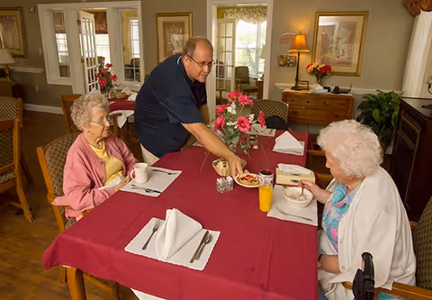 Two elderly women seated at a dining table with a red tablecloth in a well-lit room, while a man serves food to one of the women. The table is set with napkins, utensils, cups, and a glass of orange juice. The room has framed pictures on the walls, a lamp on a wooden sideboard, and floral decorations.