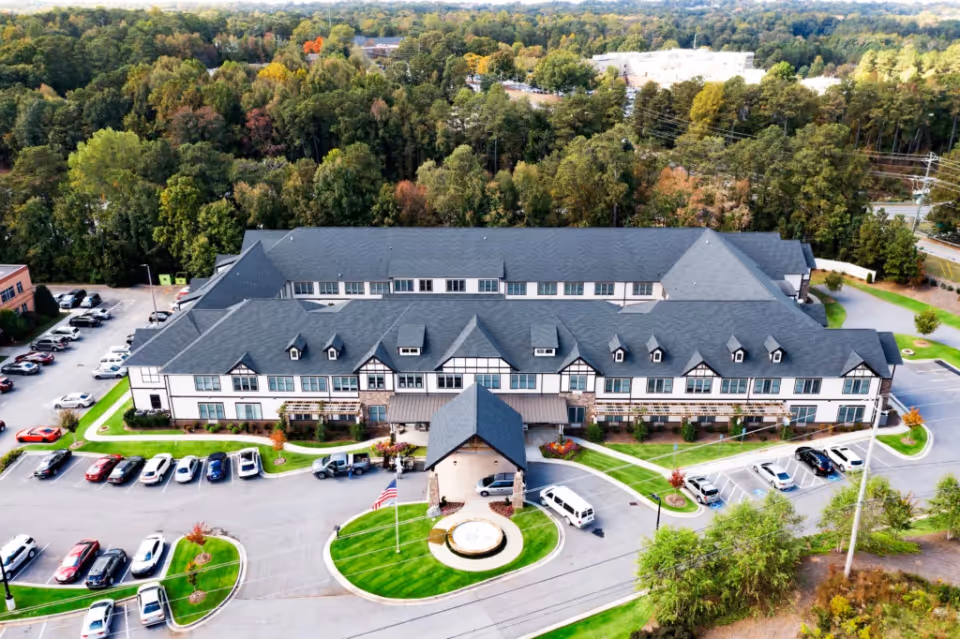 Aerial view of a large senior living facility building named Oaks at Douglasville, surrounded by parking lots with cars and green landscaped areas, with a backdrop of dense trees and a partly cloudy sky.