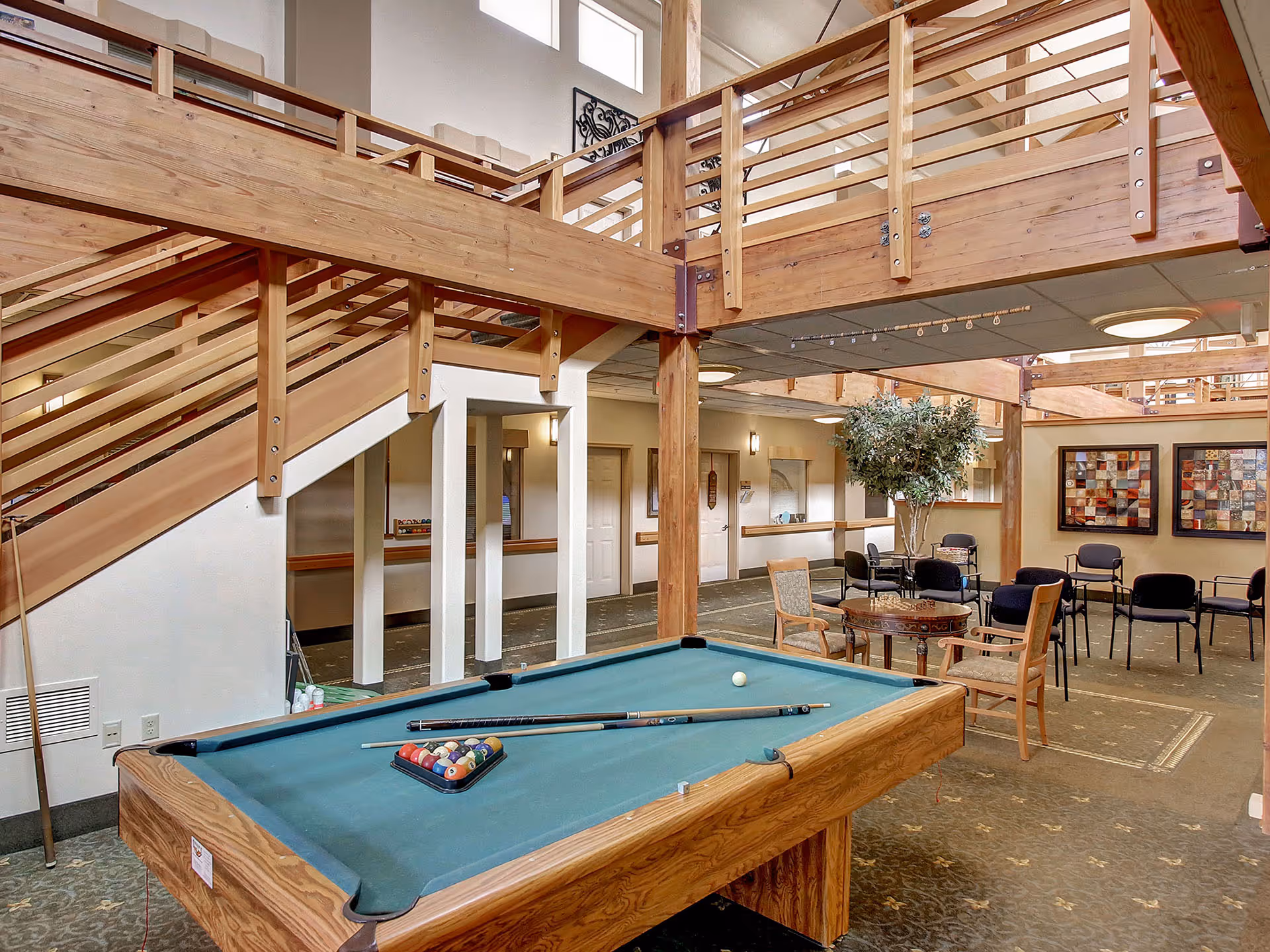 Interior view of a senior living facility common area featuring a pool table with cues and balls arranged for a game. The space includes wooden beams and railings, a staircase, several chairs and tables, decorative wall art, and a large potted plant under a ceiling with recessed lighting.