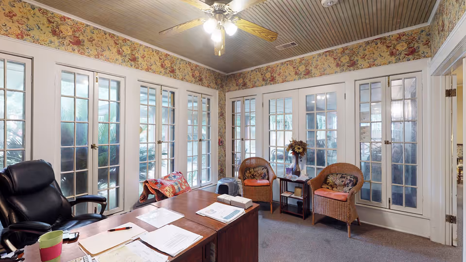 Sunlit communal living area with a desk and office chair in the foreground, wicker chairs and a side table by multiple glass-paned French doors and floral wallpaper.