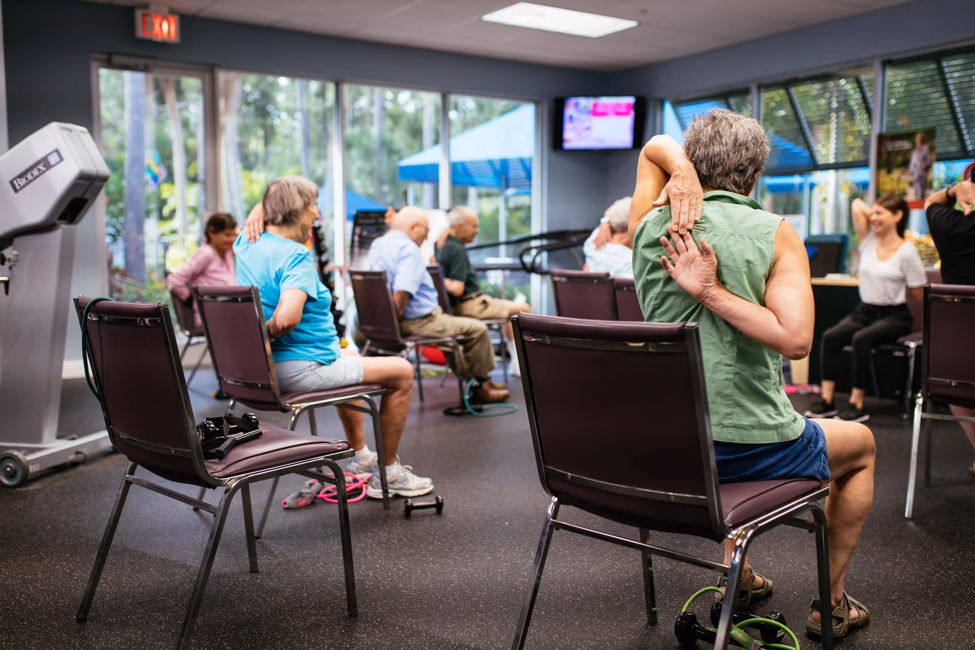 A group of elderly people sitting on chairs in a fitness room participating in a seated stretching exercise. The room has large windows showing outdoor greenery, exercise equipment, and a television mounted on the wall.