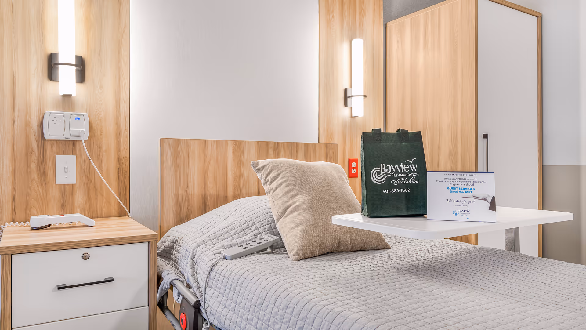 A tidy rehabilitation bedroom with a single bed, bedside table, and an overbed table holding a Bayview Rehabilitation branded bag and an information card.