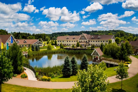 A senior living complex with beige buildings surrounding a central pond, walkways, trees, and a small gazebo under a blue sky.