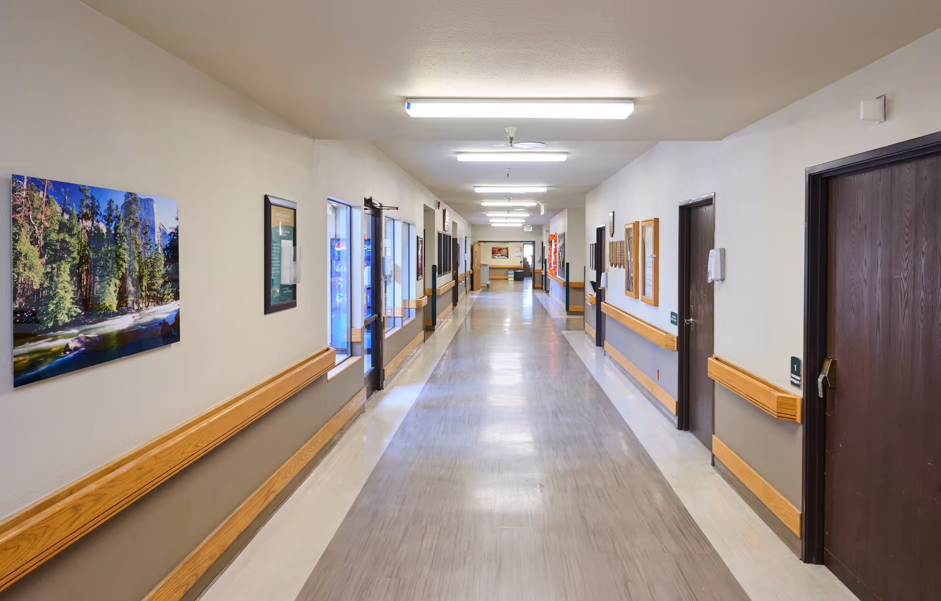 A long, clean hallway inside a nursing and rehabilitation center with beige walls, wooden handrails, several closed dark brown doors, fluorescent ceiling lights, and framed pictures and notices on the walls.