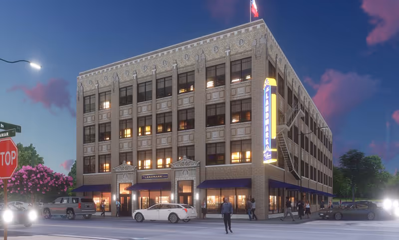 Exterior view of a four-story brick building at dusk with lit windows and a vertical illuminated sign reading 'Landmark' on the corner. Several people are walking on the sidewalks and cars are parked or driving nearby. A stop sign and street signs are visible at the intersection.