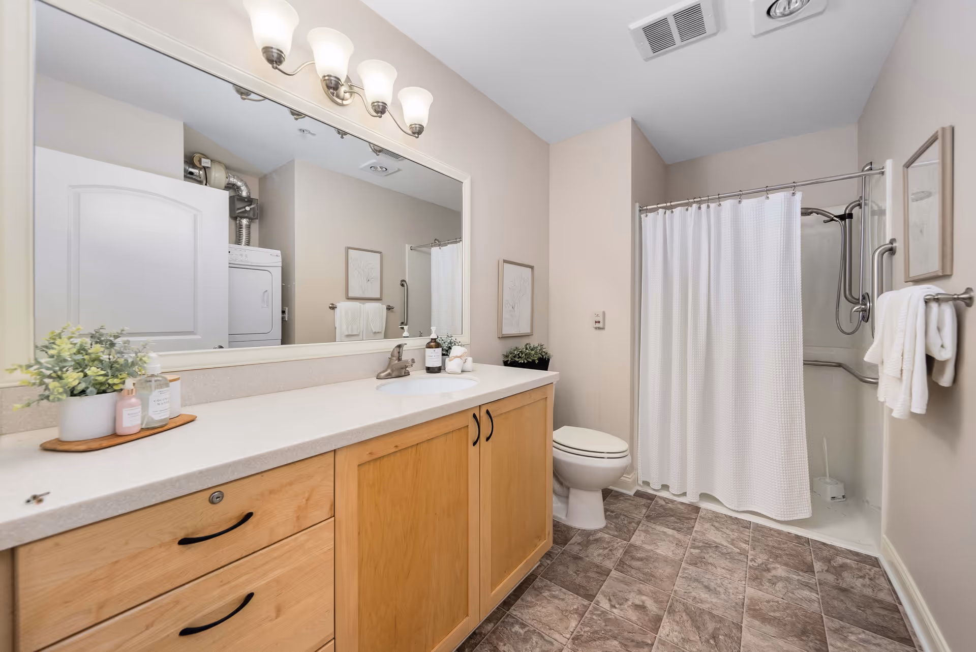A clean and well-lit bathroom featuring a large mirror above a wooden vanity with a white countertop. On the countertop, there are small decorative plants and toiletries. The bathroom includes a toilet, a shower with a white curtain, and a towel rack with white towels. The floor is covered with brown and gray tiles, and there is a laundry area with a stacked washer and dryer visible in the reflection of the mirror.