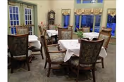 Dining area with several tables covered with white tablecloths and surrounded by upholstered wooden chairs. Large windows with beige curtains allow natural light into the room. A grandfather clock is visible in the corner.