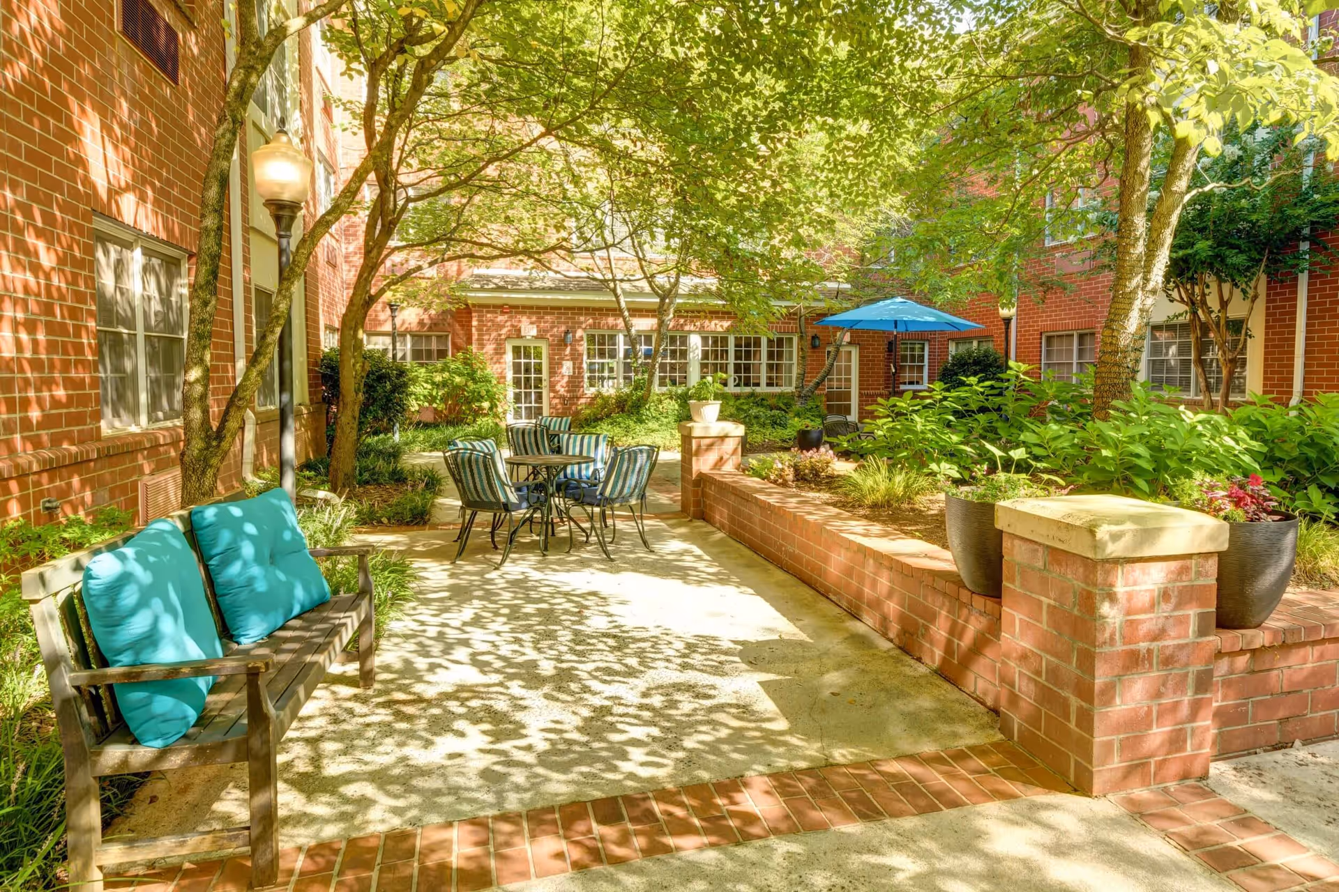 Outdoor courtyard area at Magnolia Place of Roswell featuring a wooden bench with turquoise cushions, a round table with four striped cushioned chairs, brick planters with green plants, trees providing shade, and a blue patio umbrella near a brick building with multiple windows and doors.