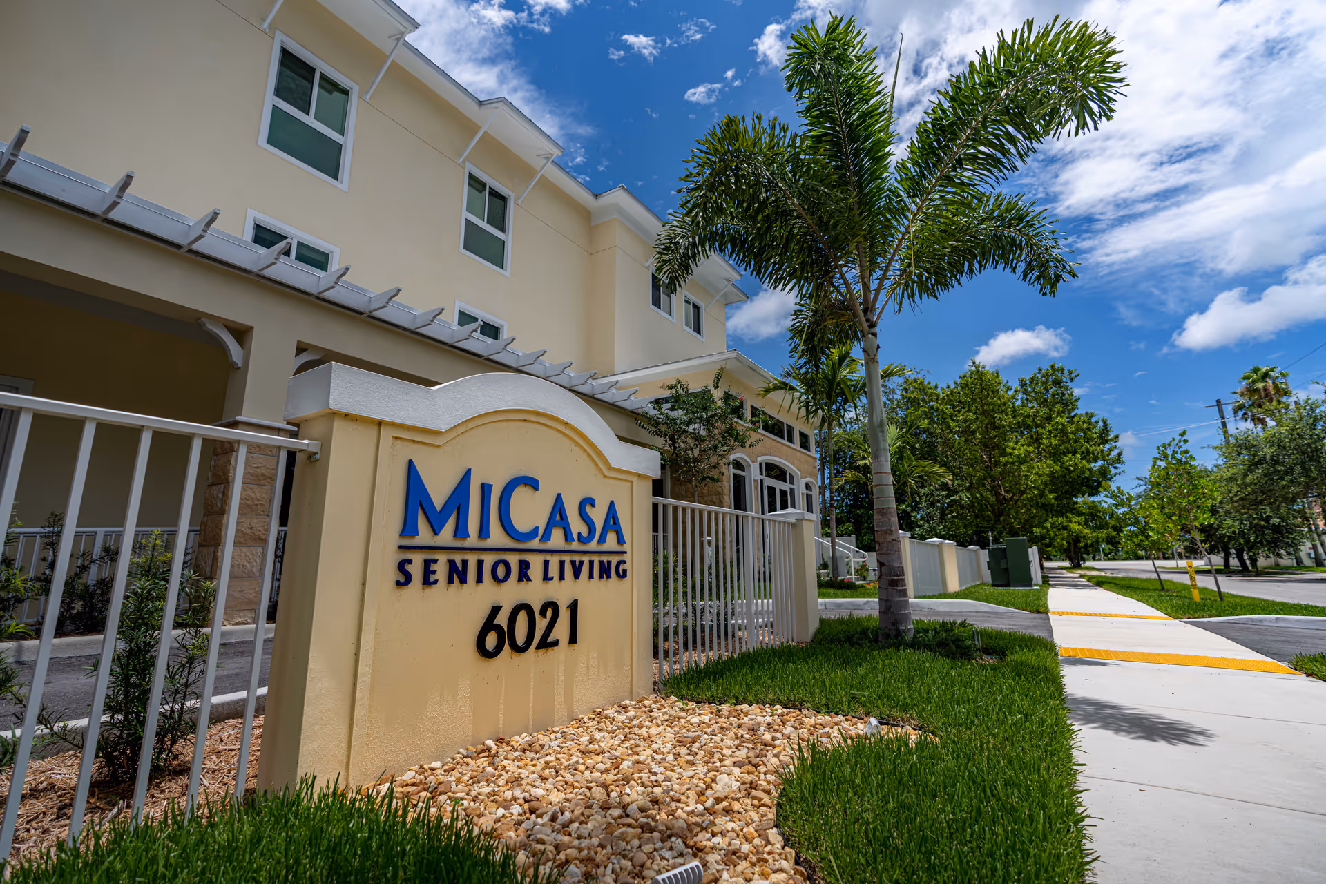 Exterior view of MiCasa Senior Living facility showing a beige building with multiple windows, a sign with the facility name and address number 6021, a palm tree, green grass, and a sidewalk under a partly cloudy blue sky.