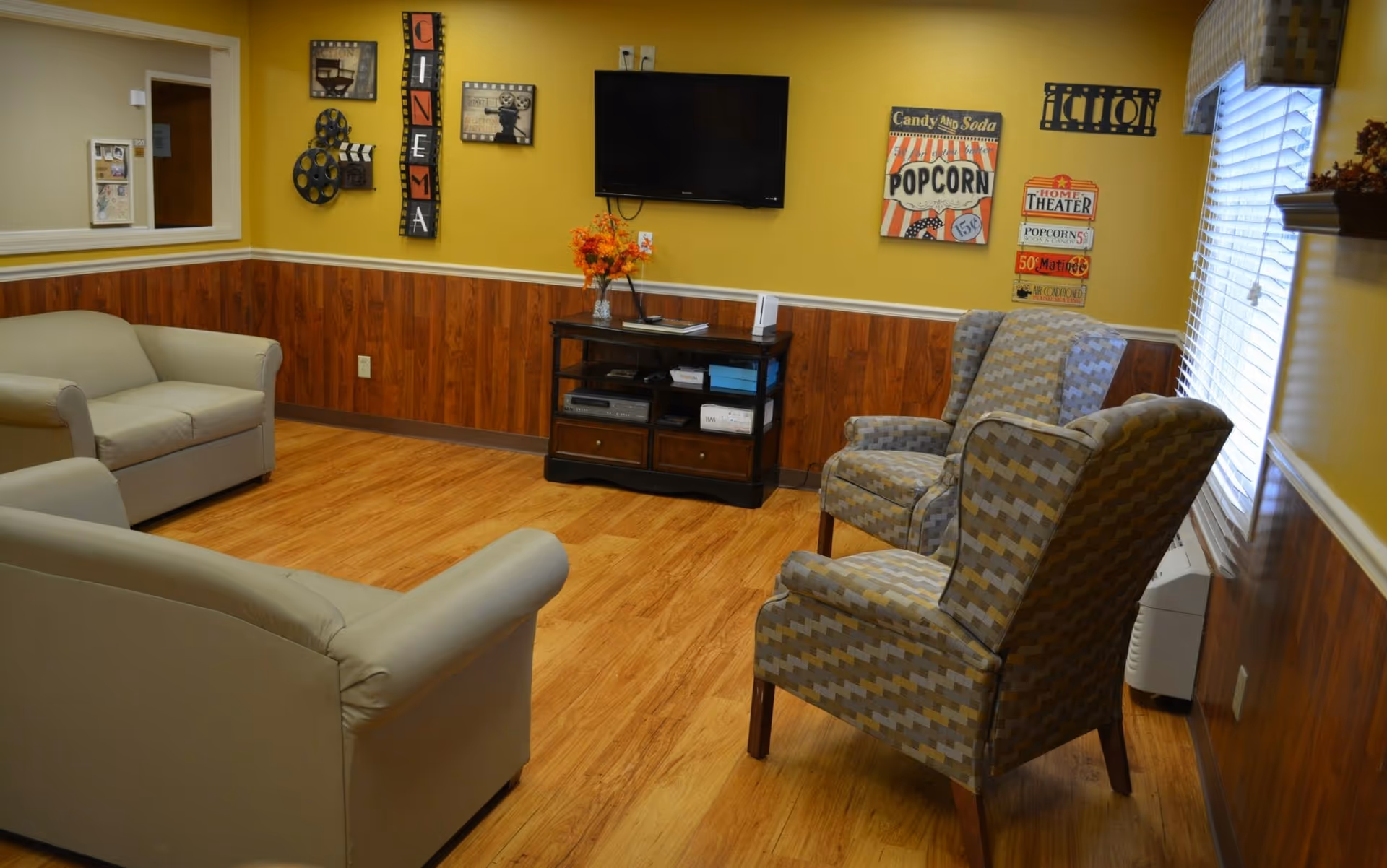 A cozy living room with two beige sofas and two patterned armchairs arranged around a wall-mounted flat screen TV. The walls are painted yellow with wood paneling on the lower half. Movie-themed decorations including signs and film reels adorn the walls. A small wooden cabinet beneath the TV holds electronic devices and a vase with orange flowers. A window with blinds and a valance is on the right side.