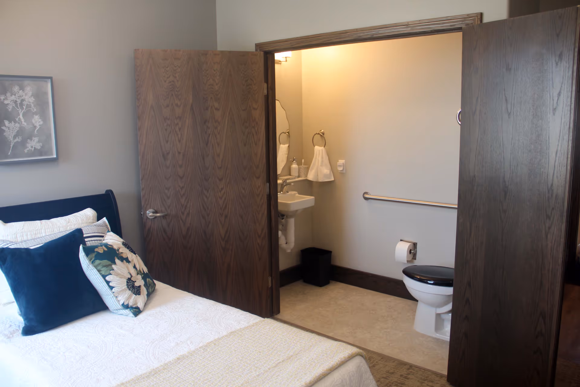 A bedroom with a made bed and decorative pillows, the door open to a small accessible bathroom with a toilet, sink, and grab bar.