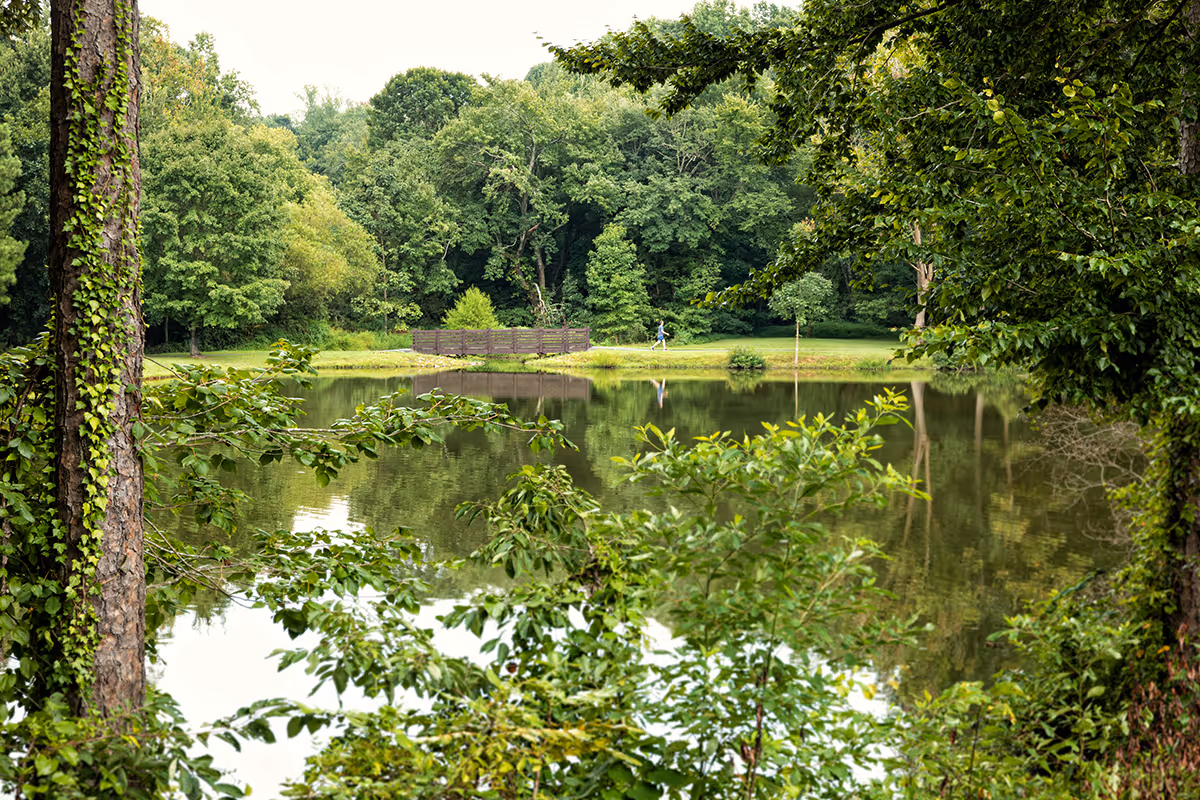 A peaceful outdoor scene featuring a small lake surrounded by lush green trees and bushes. A wooden bridge is visible across the lake, and a person is walking on a path in the distance.