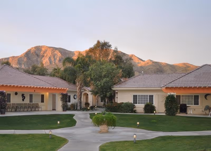 View of a senior living facility courtyard with two single-story buildings featuring tiled roofs and beige walls. The courtyard has well-maintained green lawns, a central paved walkway with a potted plant, and small pathway lights. In the background, there are tall trees and a mountain range under a clear sky during sunset.