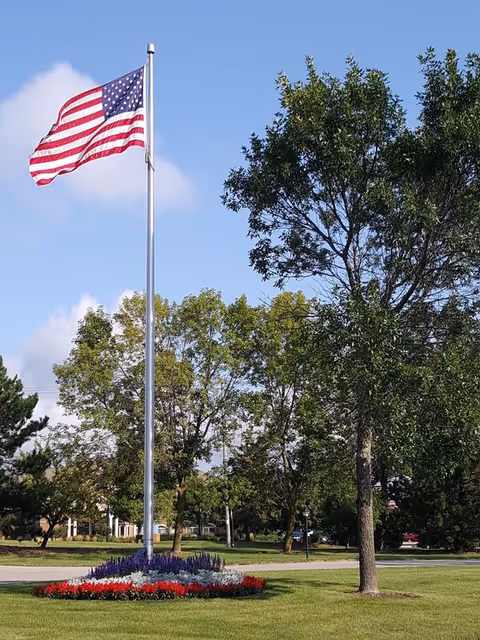 An American flag flying on a tall flagpole surrounded by a circular flower bed with red, white, and purple flowers. The flagpole is situated on a well-maintained grassy area with several trees and a clear blue sky in the background.