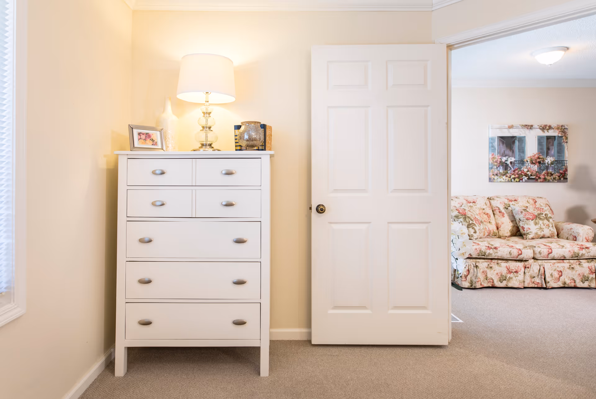 A bright room with a white chest of drawers topped with a lamp, a framed photo, and decorative items. An open white door reveals a living room area with a floral patterned sofa and a wall painting of a window with flowers.