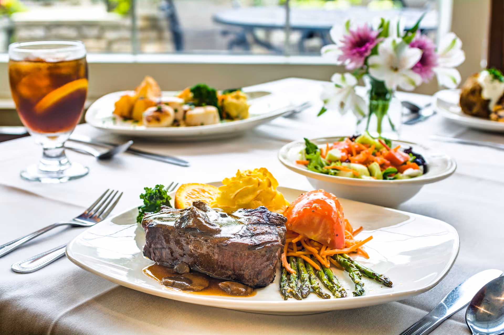 A plated meal on a white tablecloth featuring a grilled steak with mushroom sauce, mashed potatoes, asparagus, shredded carrots, and a slice of tomato. In the background, there is a bowl of salad, a glass of iced tea with a lemon slice, and another plate of food. A small vase with pink and white flowers is also visible on the table near a window.