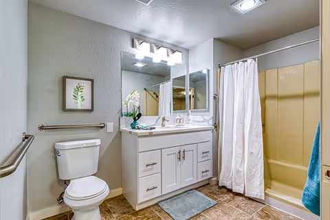 A clean and well-lit bathroom featuring a white toilet, a white vanity with a sink and multiple drawers, a large mirror with three light fixtures above it, a shower with a white curtain, and a blue bath mat on the tiled floor. There is a small framed plant artwork on the wall and a grab bar next to the toilet.