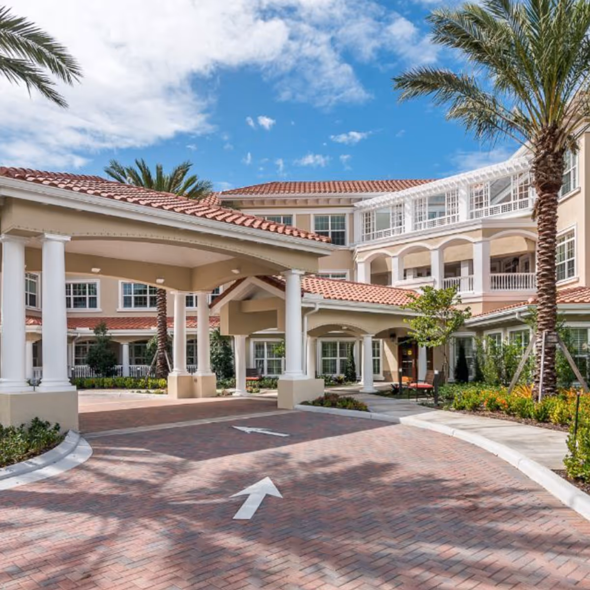 Exterior view of The Barclay at Boynton Beach senior living facility showing a covered driveway entrance with white columns, red-tiled roofs, palm trees, and landscaped greenery under a partly cloudy blue sky.