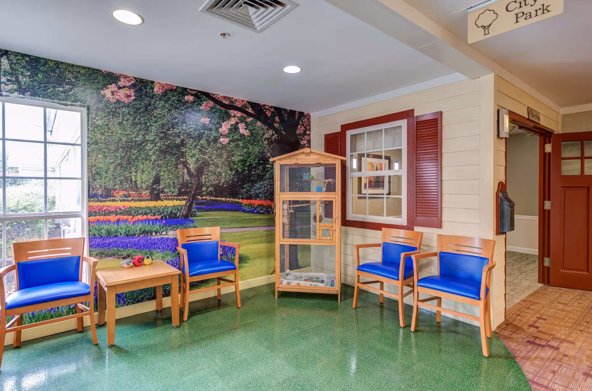 A bright seating area in a senior living facility with four wooden chairs featuring blue cushions arranged around a small wooden table. Behind the chairs is a large wall mural depicting a colorful garden with blooming flowers and trees. A wooden birdcage stands near a wall designed to look like the exterior of a house with a window and red shutters. The floor is green, and there is a hallway with a red door and a sign overhead that reads 'City Park.'