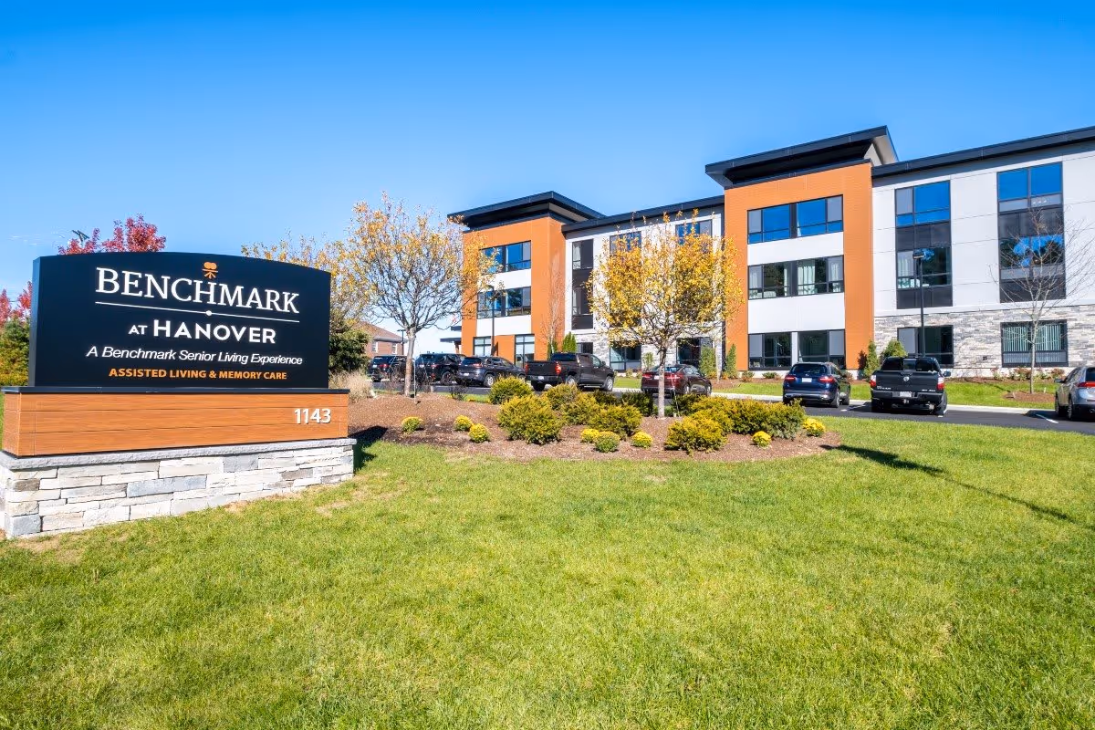 Exterior view of Benchmark at Hanover senior living facility with a large sign in the foreground displaying the facility name and description. The building is modern with multiple windows, surrounded by landscaped greenery and parked cars under a clear blue sky.