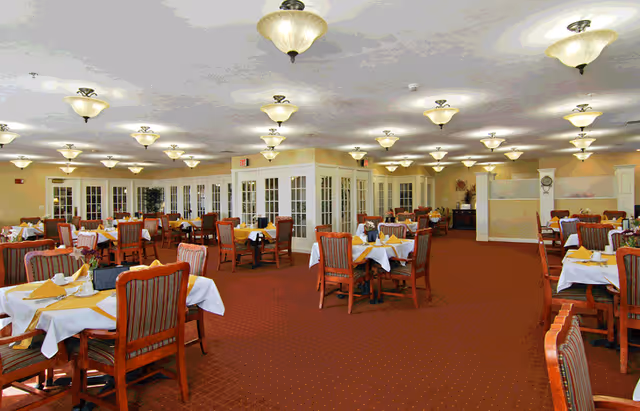 Large dining room in a senior living facility with multiple tables set with white and yellow linens and wooden chairs under many ceiling lights.