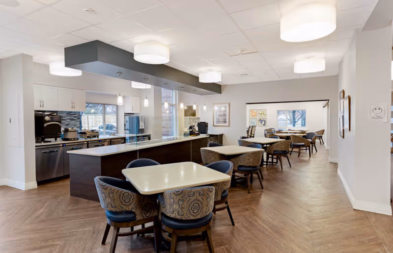 A spacious dining area in a senior living facility featuring several tables with upholstered chairs arranged neatly. The room has a modern kitchen area with stainless steel appliances and a long counter with a glass partition. The space is well-lit with ceiling lights and large windows allowing natural light to enter. The flooring is wood-patterned, and the walls are painted in light neutral colors.