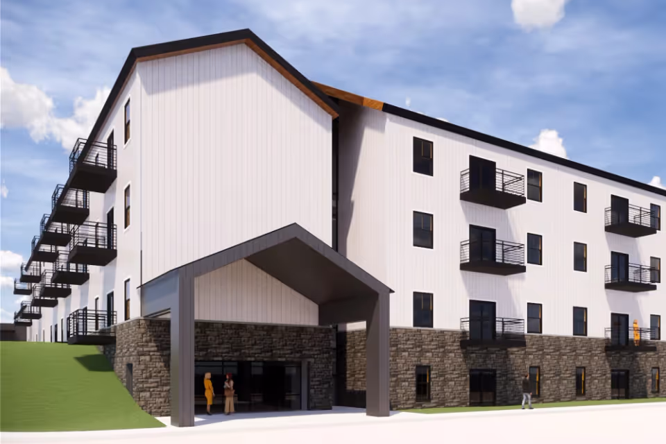 Modern four-story white apartment building with black balconies, stone-clad ground level, a covered entrance canopy, and a few people near the doorway.